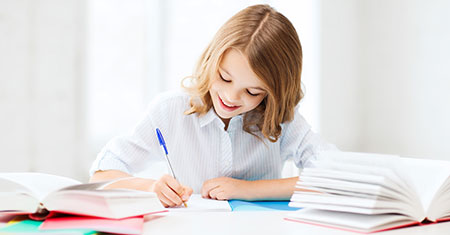 ein Mädchen sitzt umgeben von Büchern an einem Tisch und schreibt mit einem Kugelschreiber
